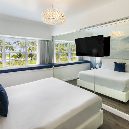 A modern bedroom with a mirrored wall, large window, two beds with white bedding and blue pillows, and a mounted TV.