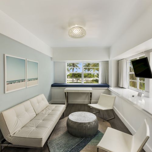 The image shows a modern, minimalist living room with a white sofa, round ottoman, two chairs, wall art, a lamp, and large windows overlooking greenery.