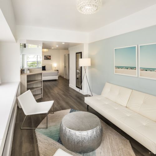 A modern living area with a white sofa, chair, round ottoman, flat-screen TV, floor lamp, and wall art featuring beach scenes. A bookshelf is in the back.