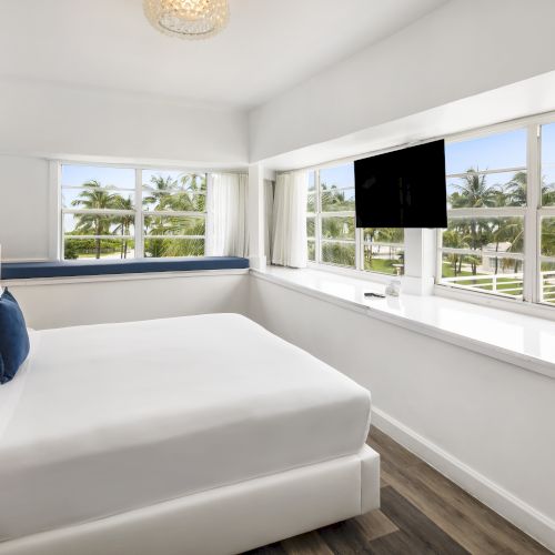 A bright bedroom with large windows, a white bed, wall art with flowers, a blue cushion bench, and a wall-mounted TV overlooking greenery outside.