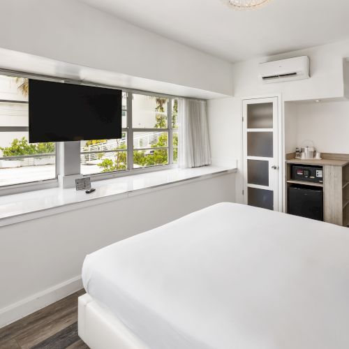 A modern hotel room with a large bed, flat-screen TV, ample windows, small kitchenette, and air conditioning unit, featuring a clean, white aesthetic.