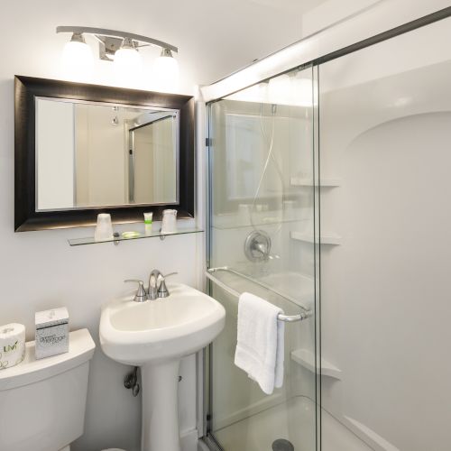 A small, modern bathroom with a shower, sink, toilet, and vanity mirror. It includes a glass shower door and a wall-mounted light fixture, ending the sentence.