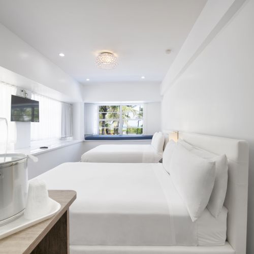 A modern hotel room with two neatly made beds, a wall-mounted TV, a refrigerated water bottle, and a window view of tropical scenery.