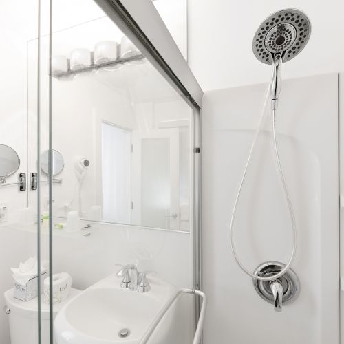 A modern bathroom with a glass shower enclosure, handheld showerhead, sink, wall-mounted hairdryer, mirror, and shelves with toiletries.