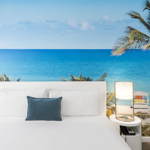 A neatly made bed with white linens and a blue pillow, framed by two lamps, set against a wall mural of an ocean view with palm trees ends the sentence.