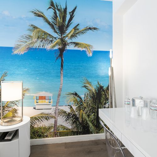 A room with white furniture has a wall mural of a beach scene, including a palm tree and blue ocean, and a photo frame.