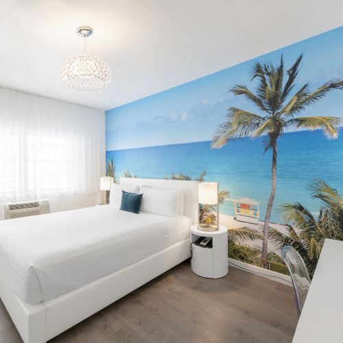 A modern bedroom features a beach-themed mural, white furnishings, a large bed, nightstands, lamps, a chandelier, a window with sheer curtains, and a desk.