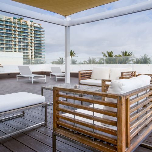 The image shows an outdoor patio with modern wooden furniture, white cushions, and loungers, set against a backdrop of palm trees and buildings.