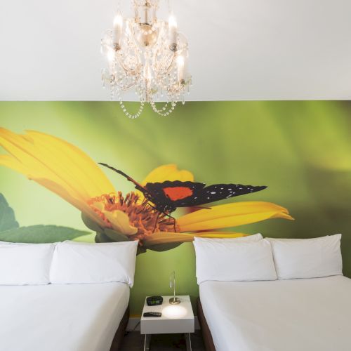 The image shows a hotel room with two white beds, a small nightstand, a chandelier, and a large wall mural of a butterfly on a yellow flower.