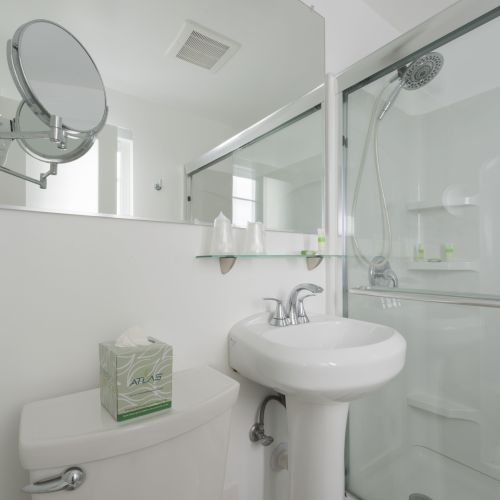 A clean, white bathroom with a sink, a mirror, a tissue box on a toilet, and a glass-enclosed shower with a showerhead and shelf.