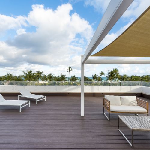 The image shows a spacious outdoor deck with lounge chairs, a shaded area, a sofa, and a table, overlooking a scenic view with palm trees and clouds in the sky.