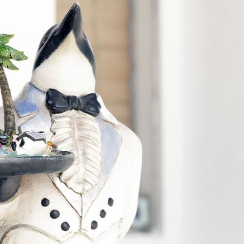 The image shows a penguin statue dressed in a tuxedo, holding a tray with a small palm tree and various miniature items on it.