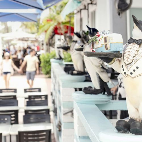 The image shows a row of penguin statues dressed as waiters, each holding a dish, at an outdoor restaurant with people walking in the background.