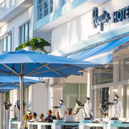 This image shows the exterior of a hotel with a blue awning and outdoor seating area occupied by people beneath blue umbrellas, labeled 