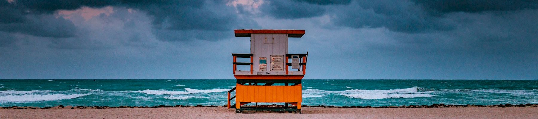 A lifeguard tower stands alone on a sandy beach with dark, dramatic clouds looming over a turbulent sea in the background.