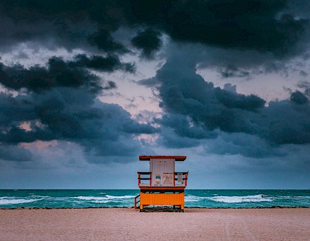 A lifeguard tower stands alone on a sandy beach with dark, dramatic clouds looming over a turbulent sea in the background.