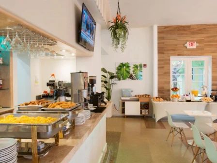 The image shows a bright cafeteria or breakfast room with food serving stations, tables, chairs, and a TV on the wall ending the sentence.