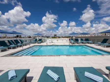 A rooftop swimming pool with lounge chairs and umbrellas under a blue sky with fluffy clouds. Towels are neatly placed on the lounge chairs.