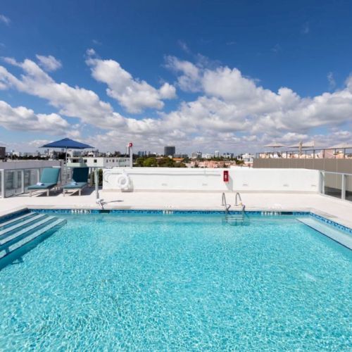 The image shows a rooftop pool with clear blue water, surrounded by lounge chairs and umbrellas. The sky is partly cloudy with blue patches.