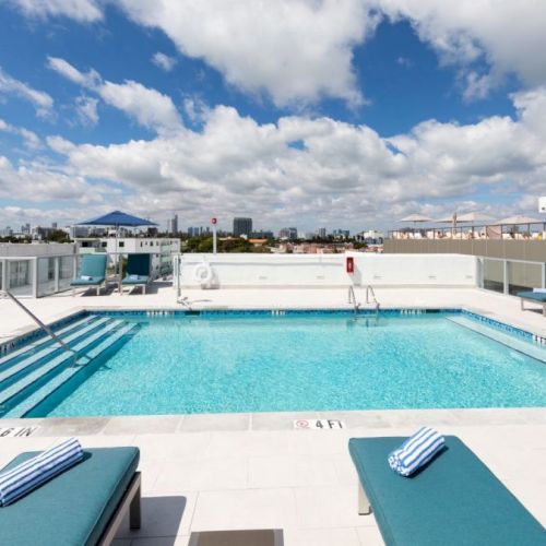 A rooftop swimming pool with lounge chairs, striped towels, and umbrellas. The sky is partly cloudy. A cityscape is visible in the background.